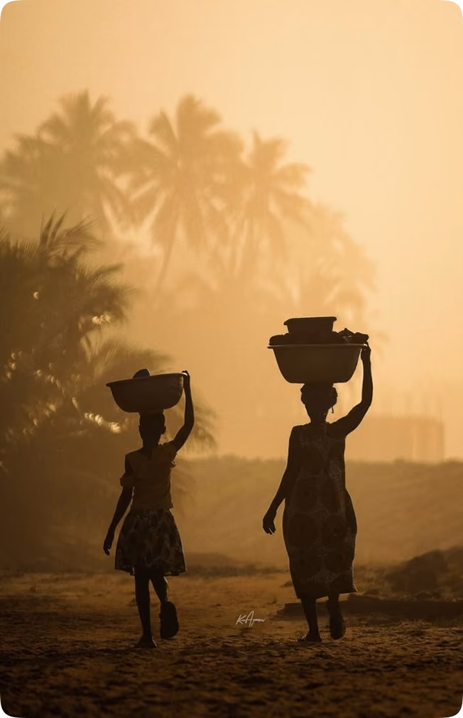Silhouettes of two women walking with baskets on their heads against a backdrop of palm trees at sunset.