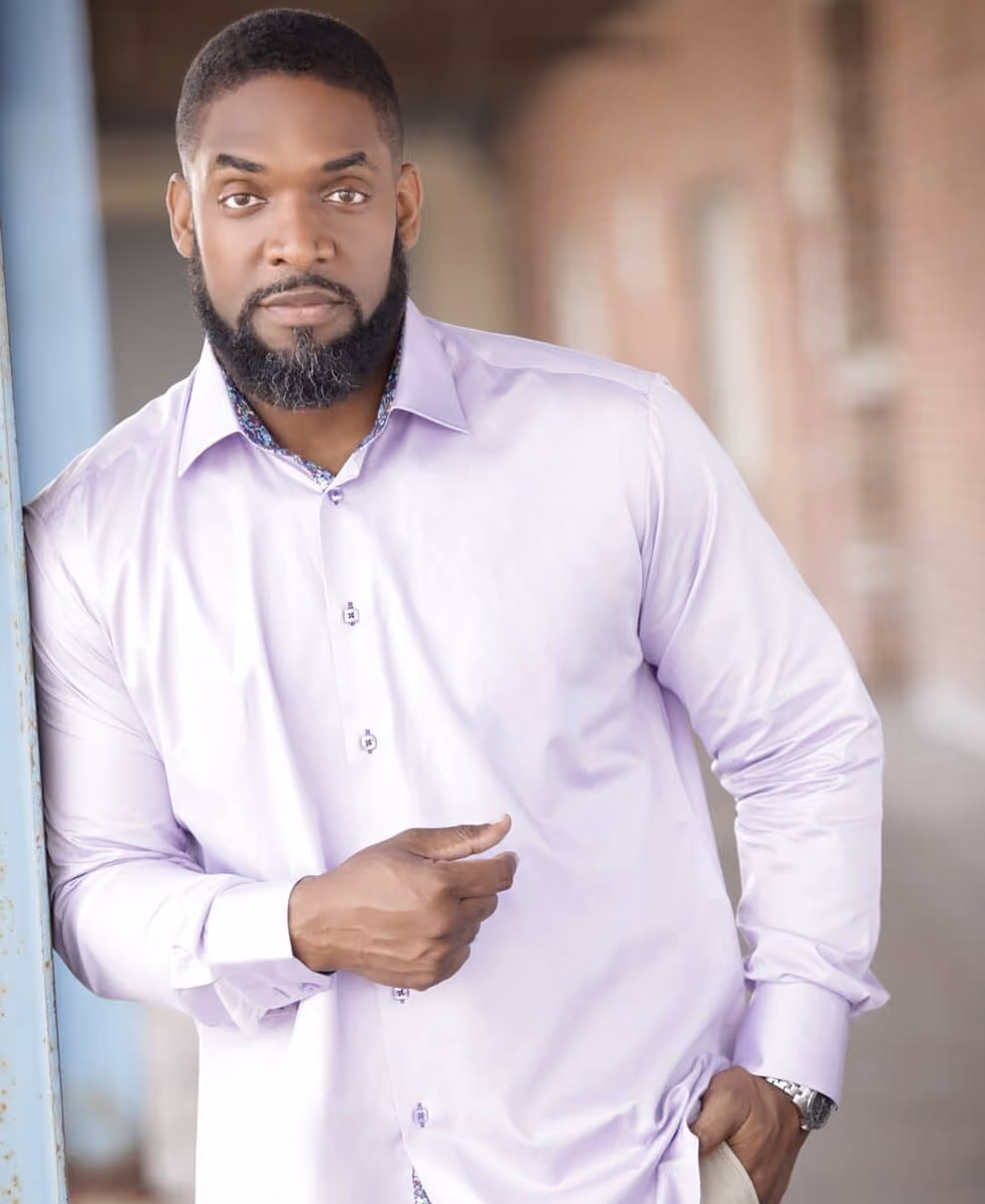Man with beard in light purple dress shirt leaning against a pole, looking at camera in a blurred corridor.