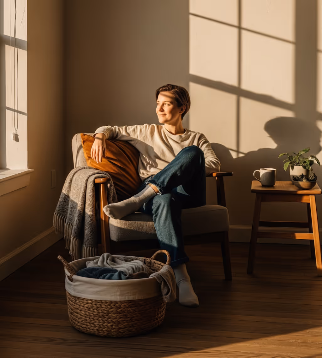Persona sentada en un sillón junto a una ventana, con luz natural, una mesa pequeña con planta y taza y una cesta con ropa en el suelo.