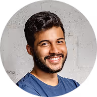 Hombre joven sonriendo con cabello rizado y barba corta, vestido con camiseta azul, frente a una pared de concreto gris.