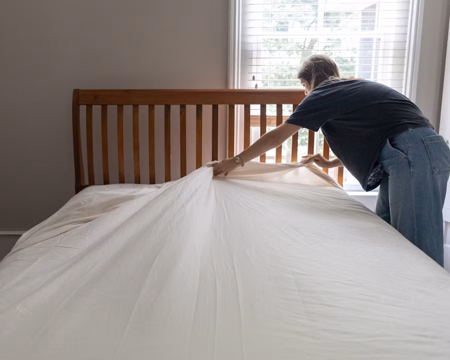 Persona haciendo la cama estirando una sábana blanca en un dormitorio con ventana y cabecera de madera.