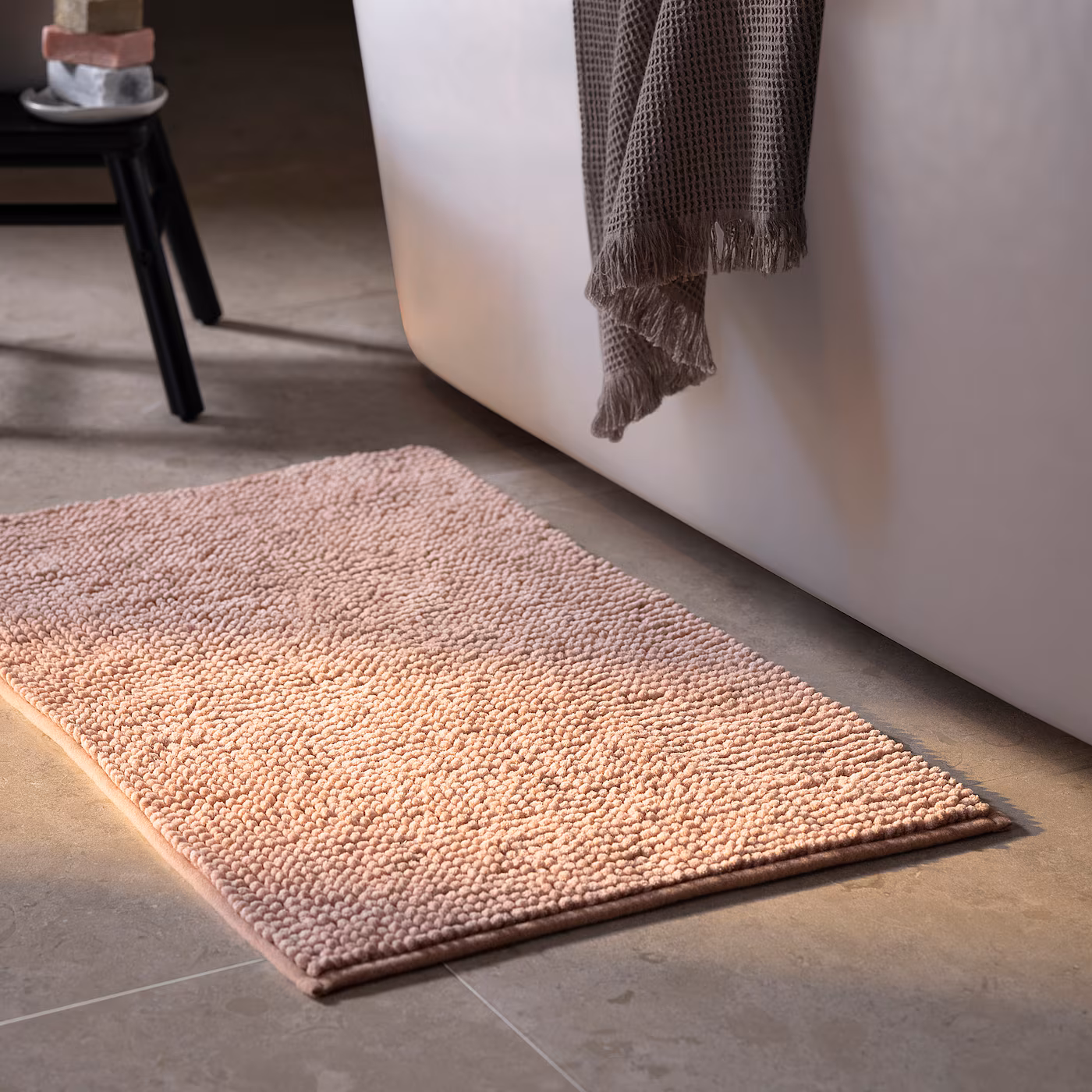 Tapete de baño rosa claro con textura de borlas frente a una bañera blanca con una toalla gris colgada.