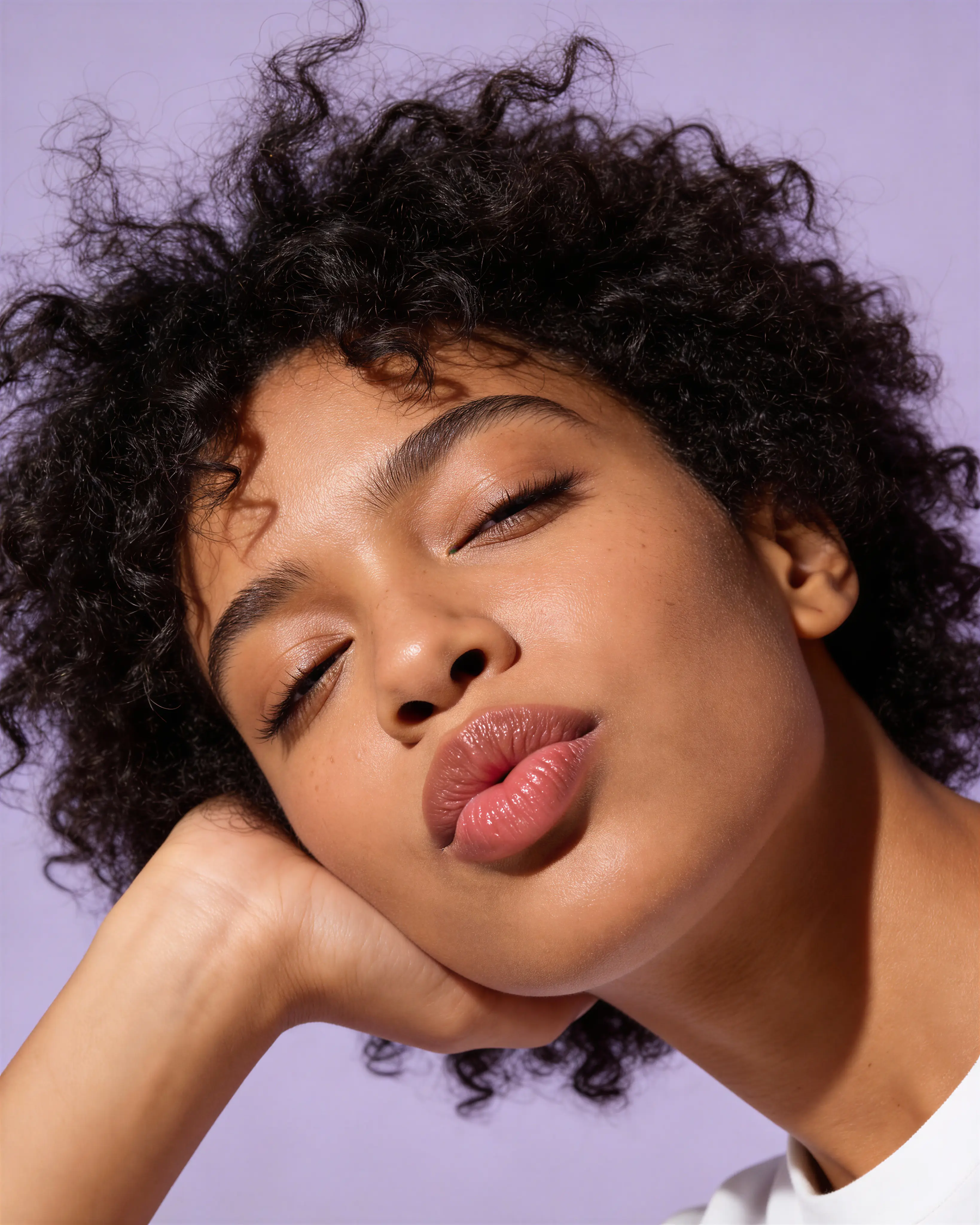 Close-up of a person with curly hair resting their head on their hand with eyes slightly closed and lips puckered against a purple background.