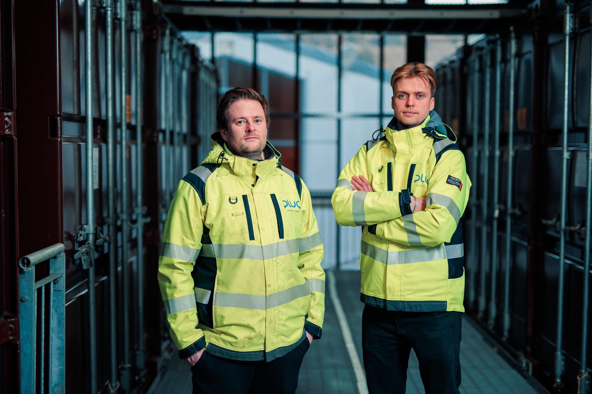 Two men in yellow high visibility jackets standing in an industrial corridor with metal containers on both sides.