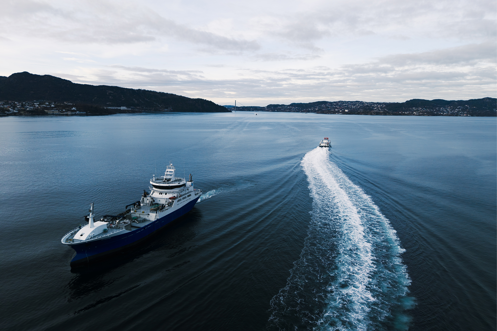 Large ship and small boat moving on calm water near a mountainous coastline under a cloudy sky.
