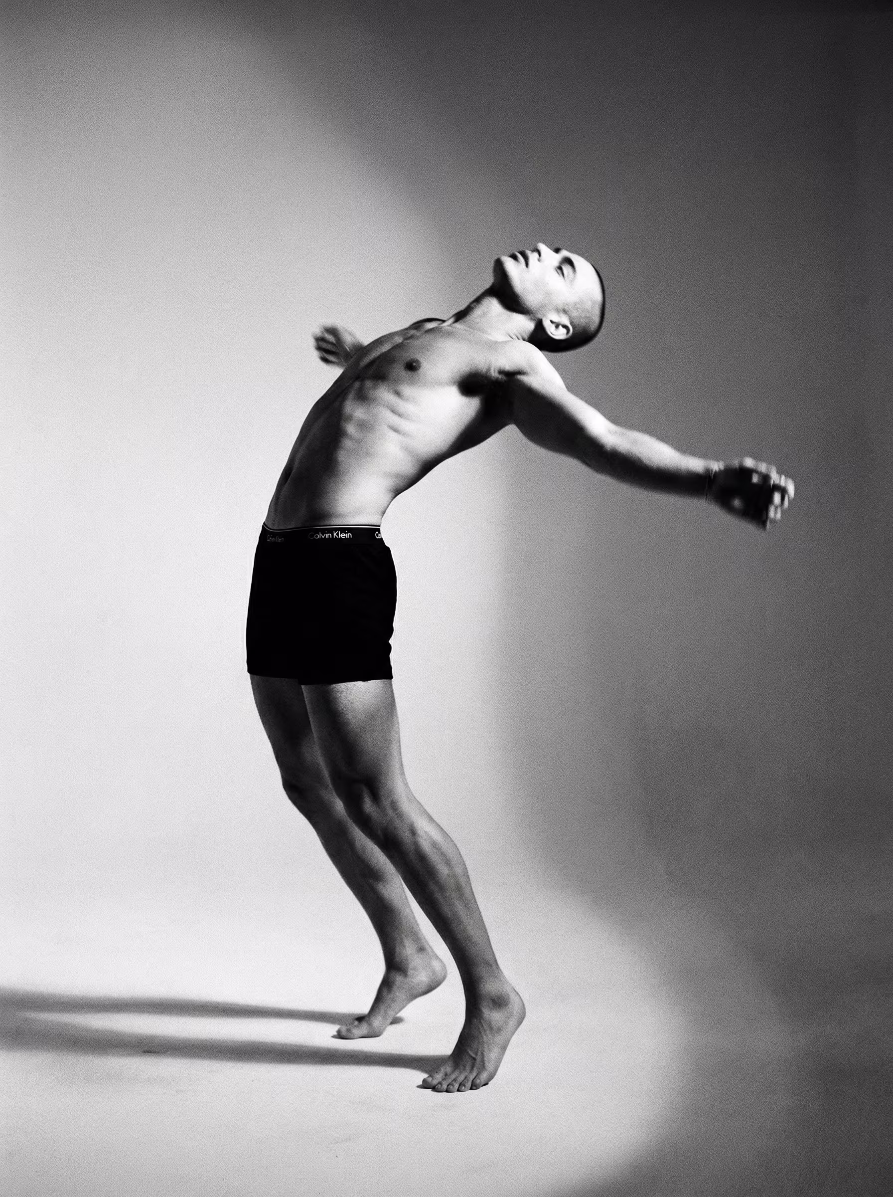 Black and white photo of a shirtless man arching back with arms extended wearing black Calvin Klein underwear.