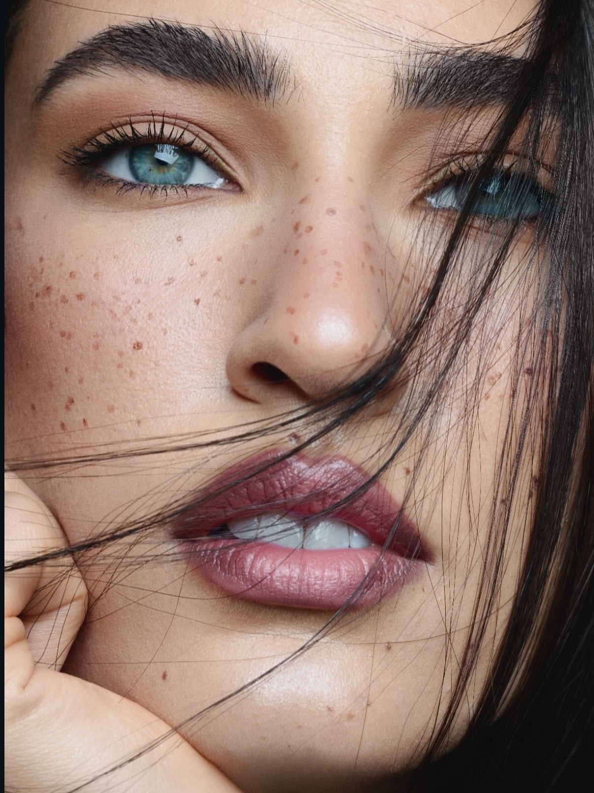 Close-up of a freckled woman with closed eyes lying on sand near orange lilies.