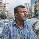 Middle-aged man in a checkered shirt standing outdoors with buildings and cars in the background.