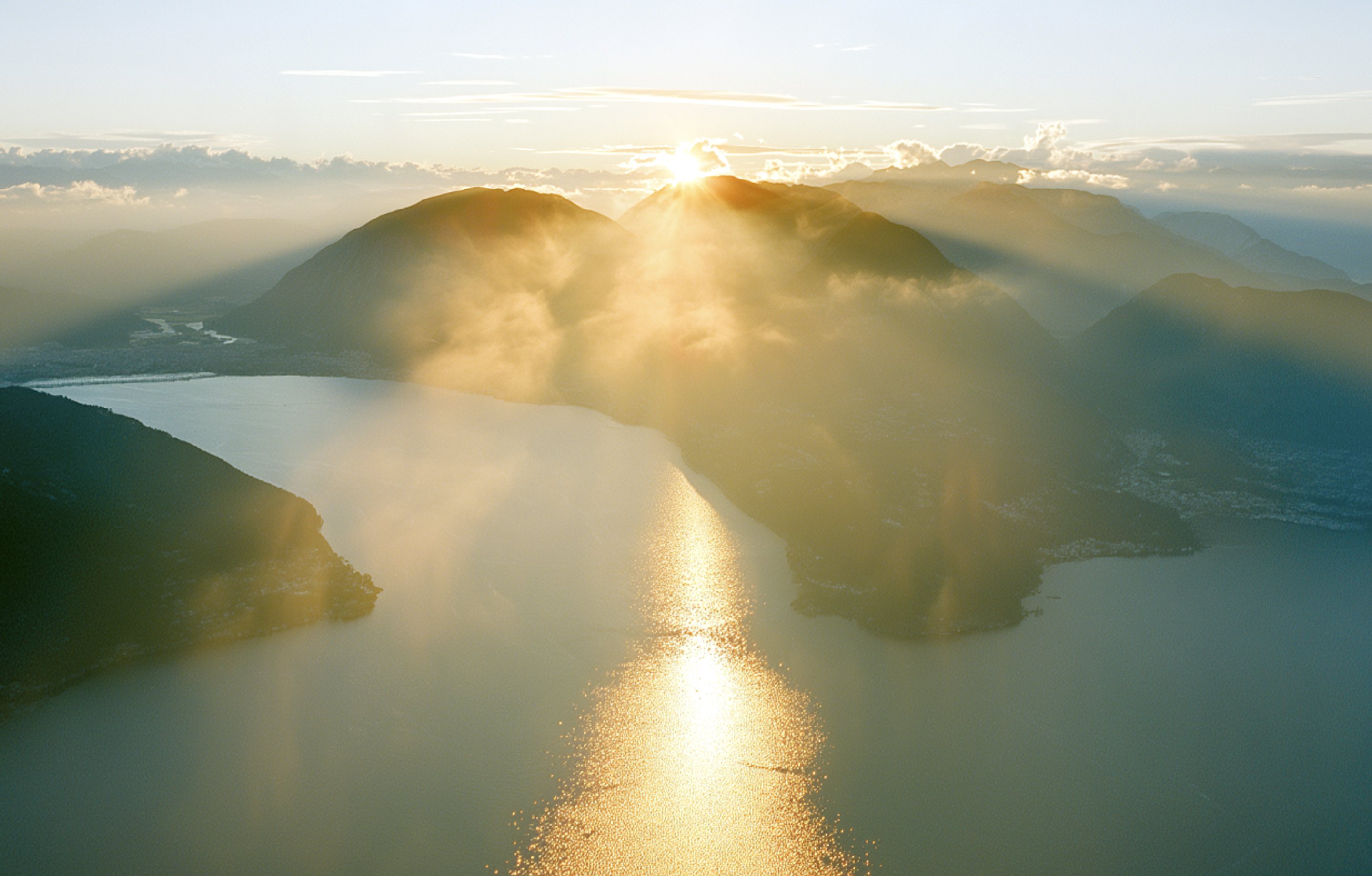 First light over Lake Lugano: the sun breaking over the mountains and a path of light on the water