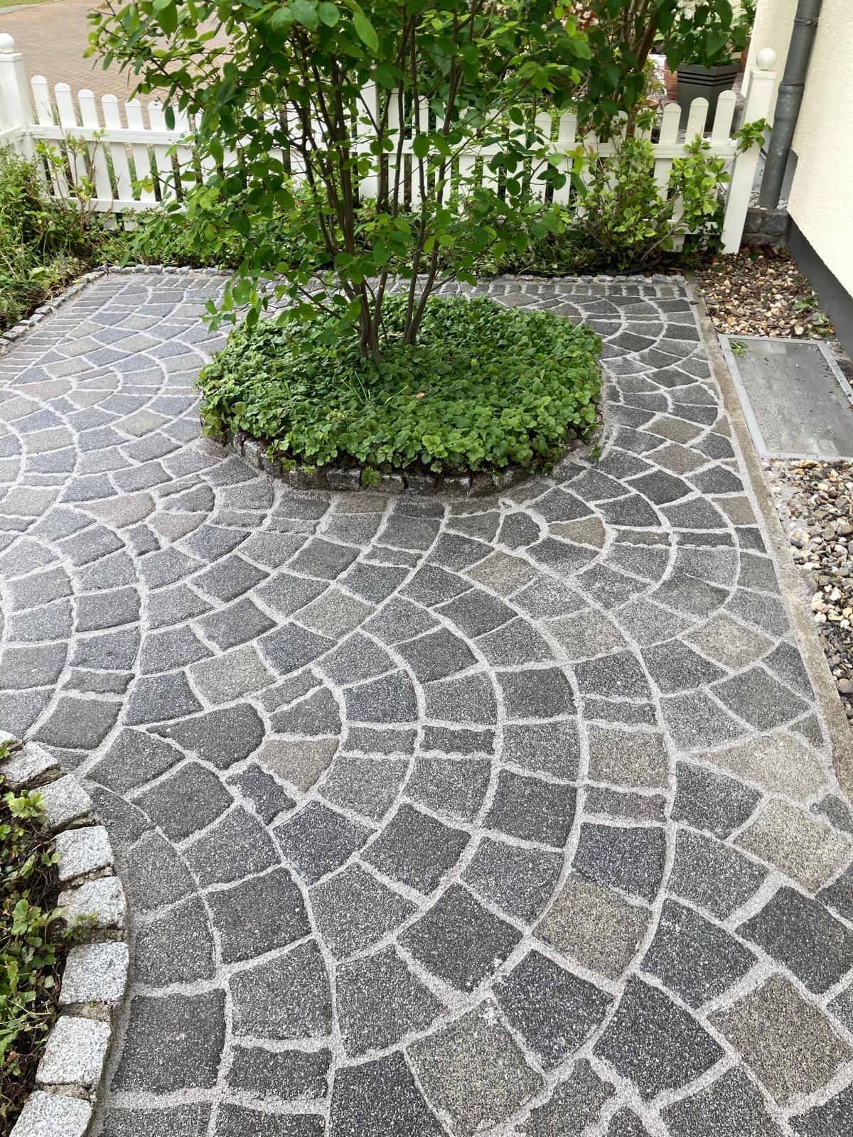 Curved gray stone patio with circular garden bed and green shrub in front of white picket fence.