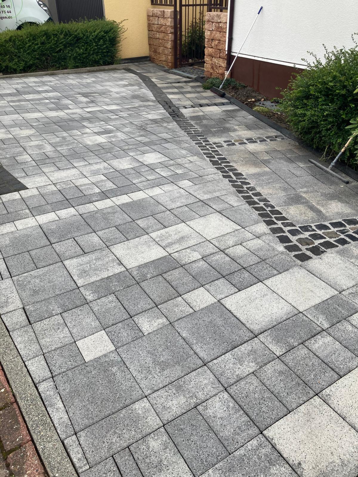 Gray and white patterned stone tile driveway with black stone border and garden tools nearby.