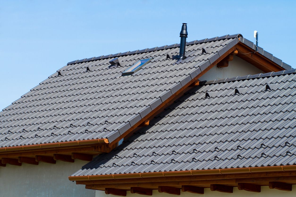 House roof with gray tiles, skylight window, pipe, and wooden eaves under clear blue sky.