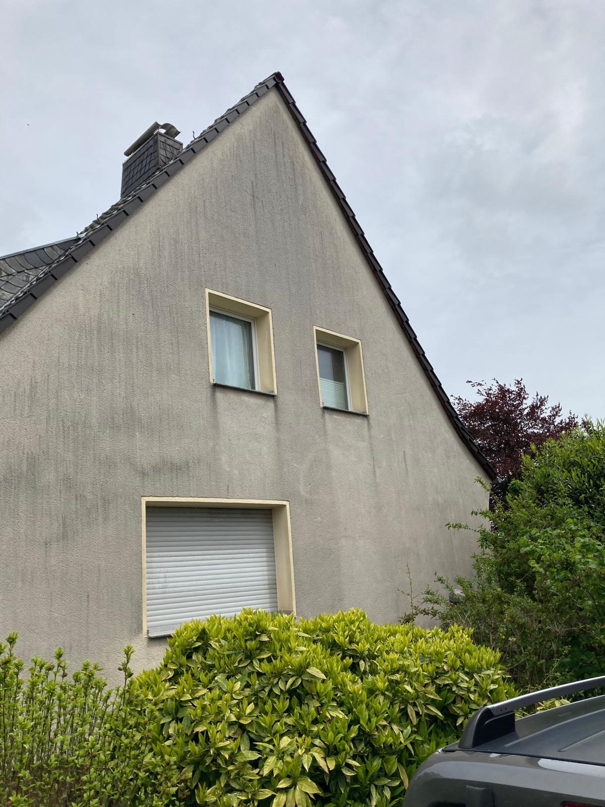 Side of a house with two upper windows, one lower closed window, green bushes, and a car roof visible.
