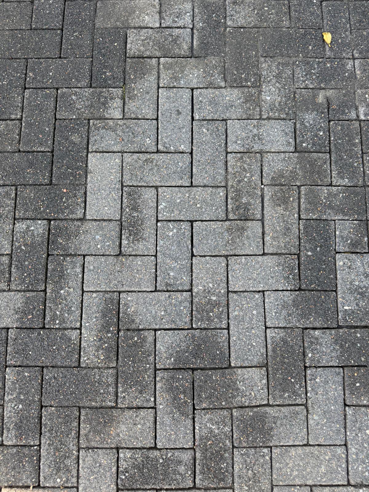 Grey interlocking rectangular bricks laid in a herringbone pattern on pavement with a small yellow leaf.