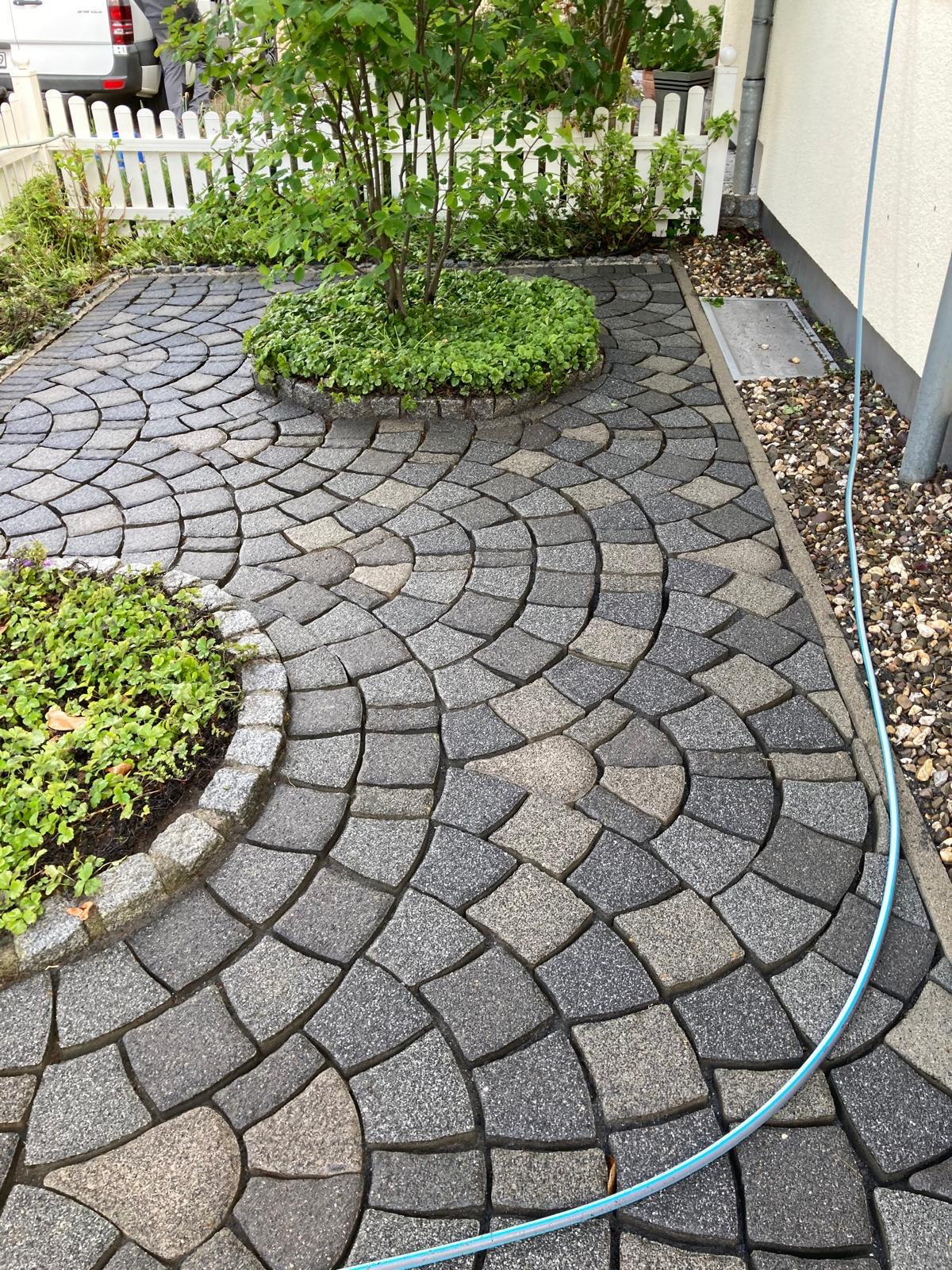 Curved stone paver patio with two round planting beds and a blue garden hose along edge.