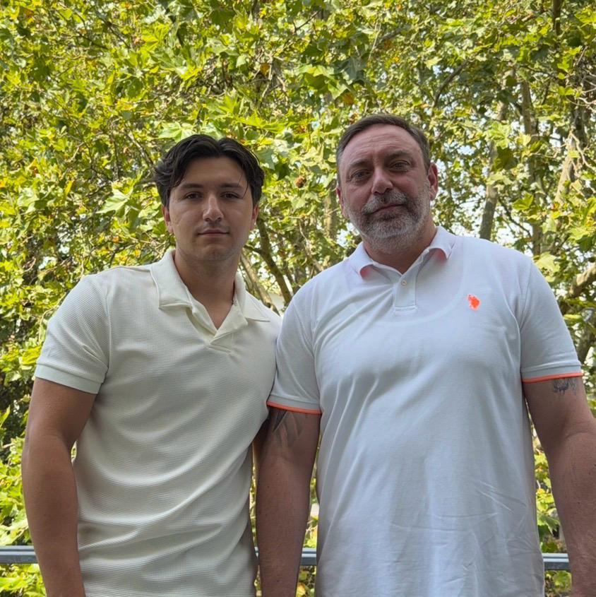 Two men standing outdoors in light-colored polo shirts with trees in the background.