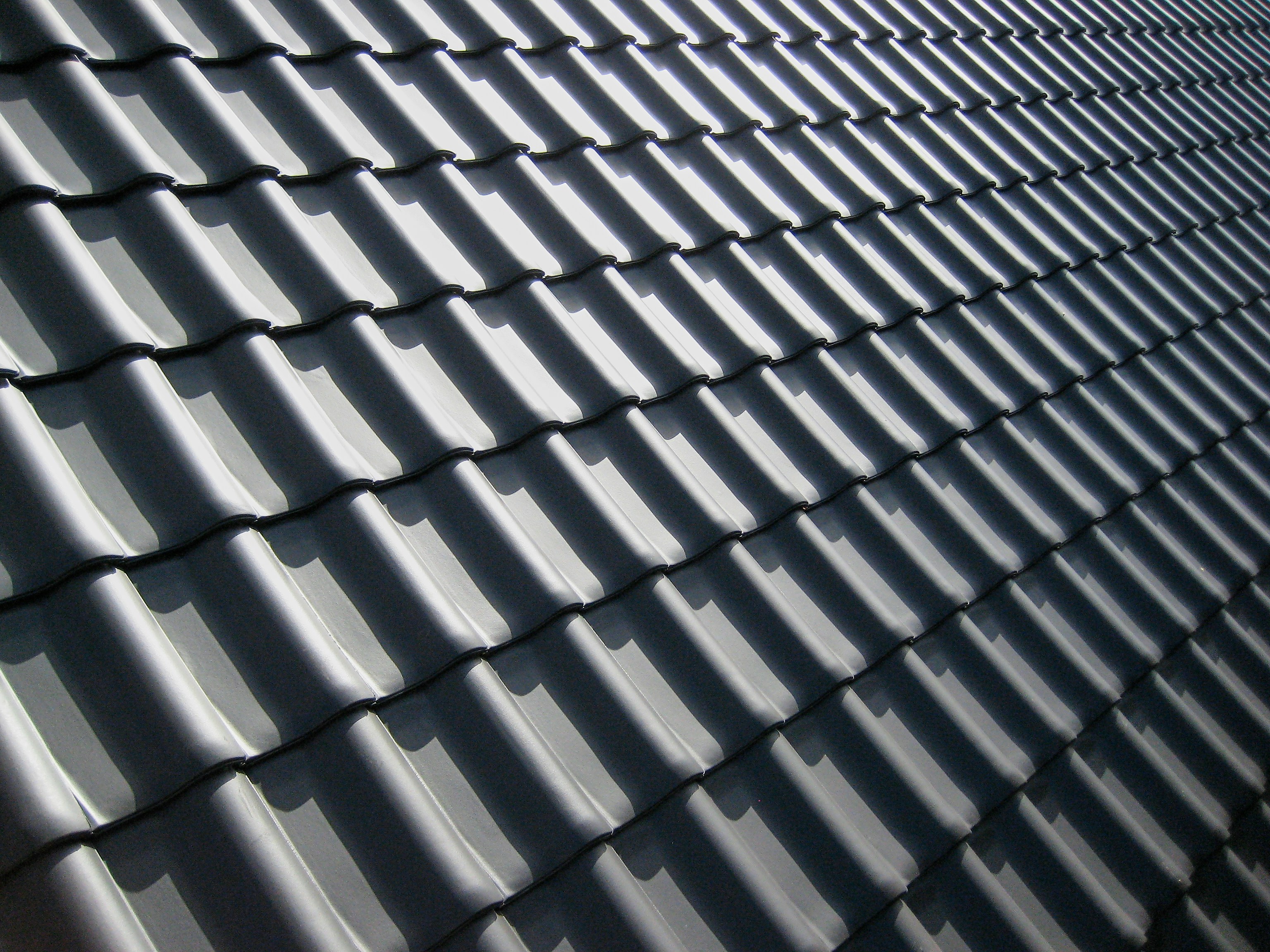 Close-up of a dark gray metal roof with a repeating pattern of ridged tiles casting shadows under sunlight.