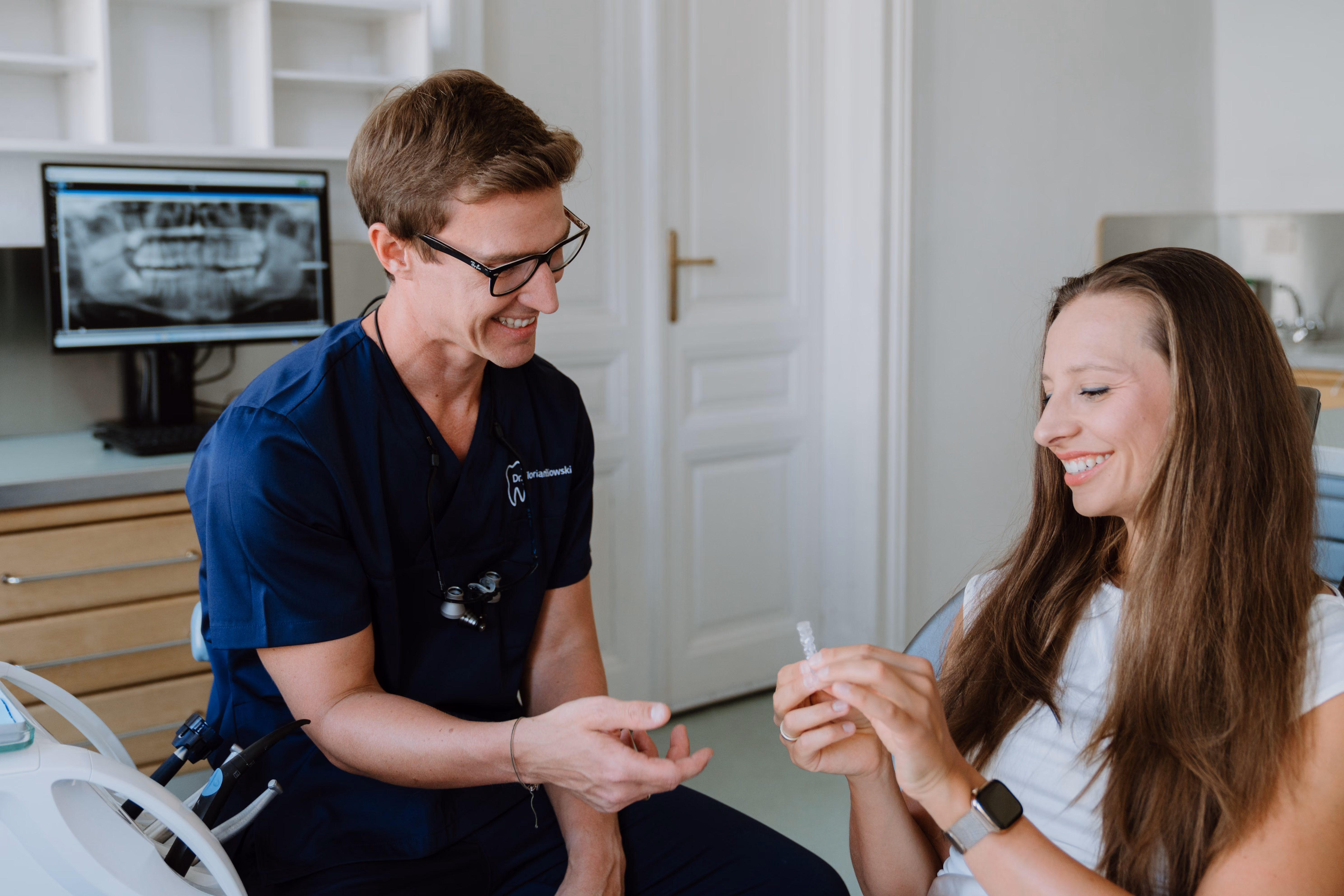 Zahnmediziner erklärt einer lächelnden Patientin in der Praxis eine durchsichtige Zahnspange.
