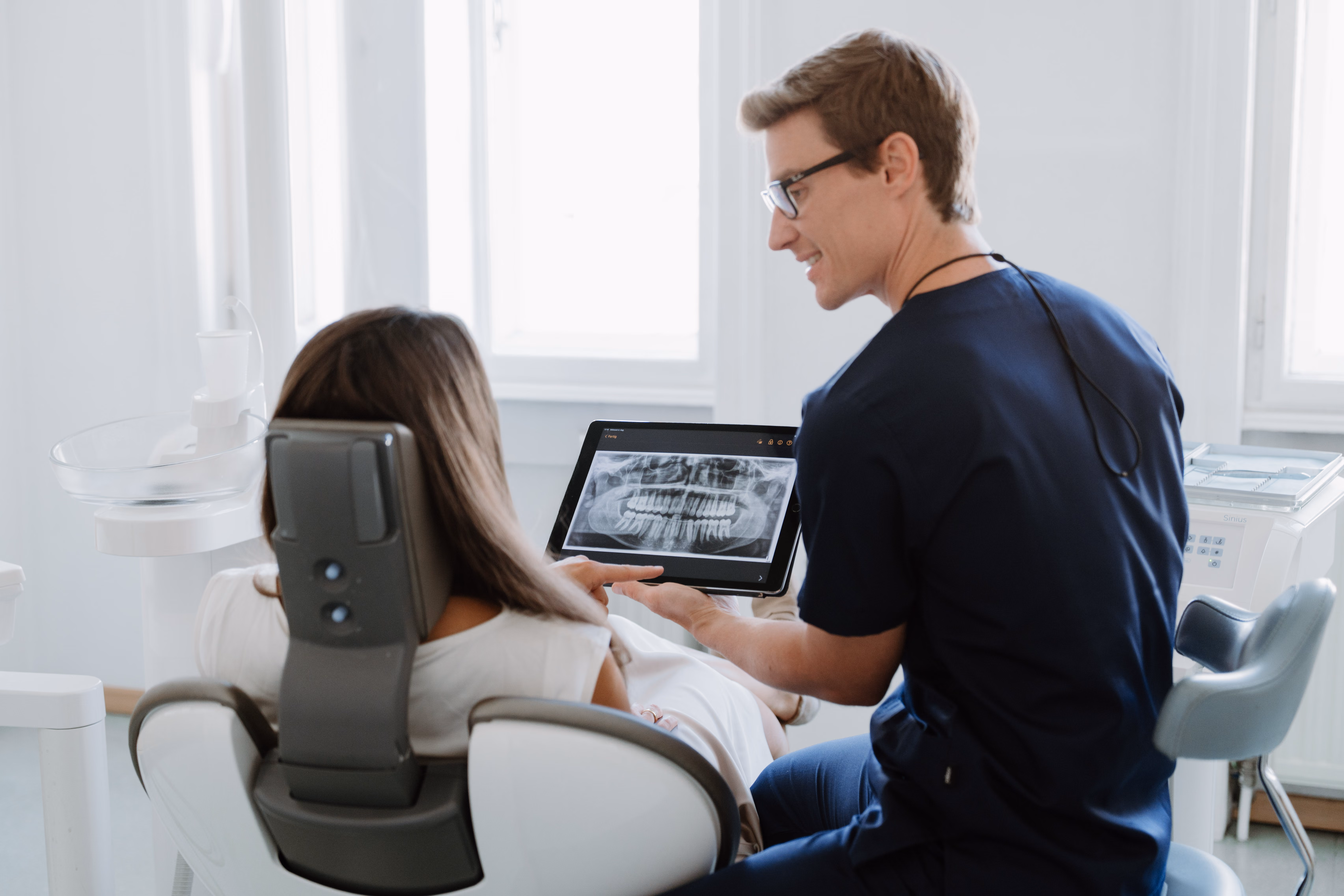 Zahnarzt zeigt Patientin ein digitales Zahn-Röntgenbild auf einem Tablet im Behandlungszimmer.