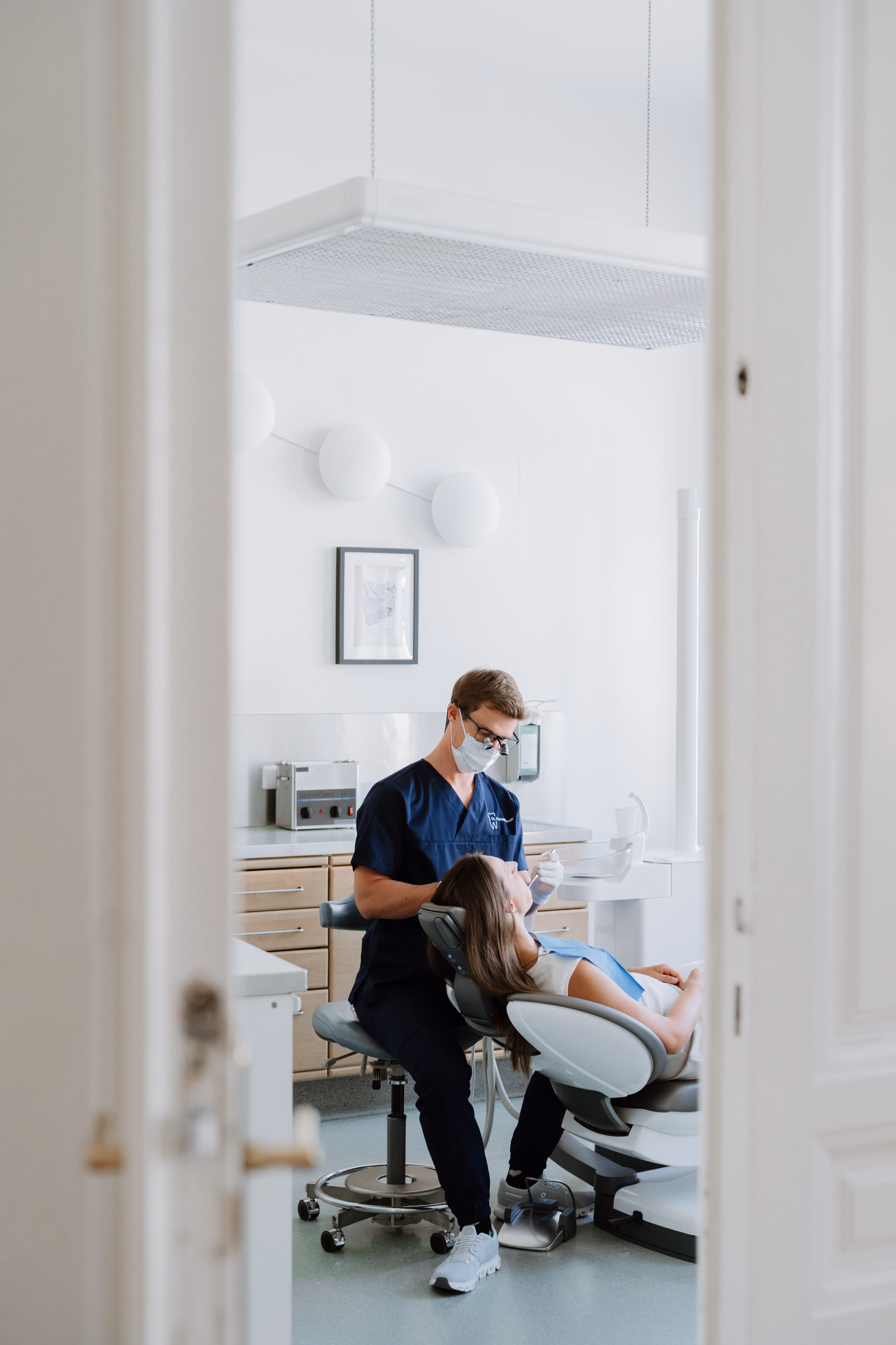 Zahnarzt mit Maske behandelt sitzende Patientin in heller, moderner Praxis.