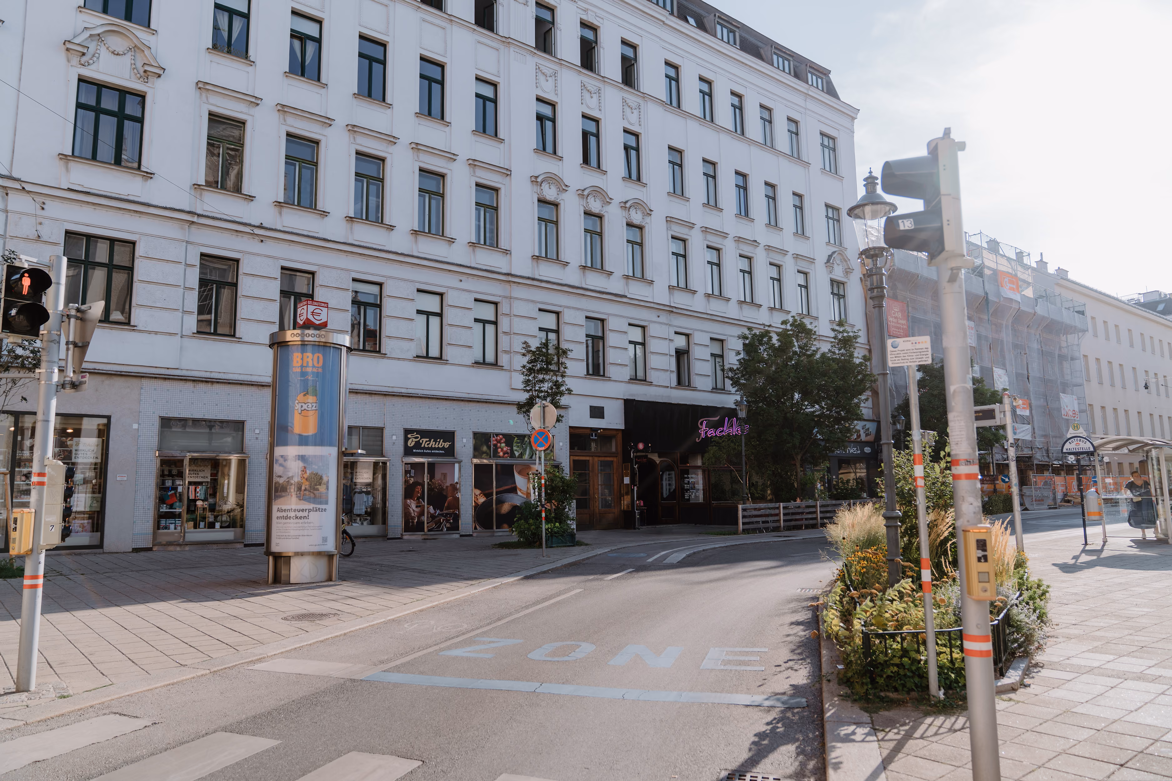 Straße mit Zebrastreifen, Altbau mit Geschäften, Ampeln und Werbesäule im Sonnenlicht.