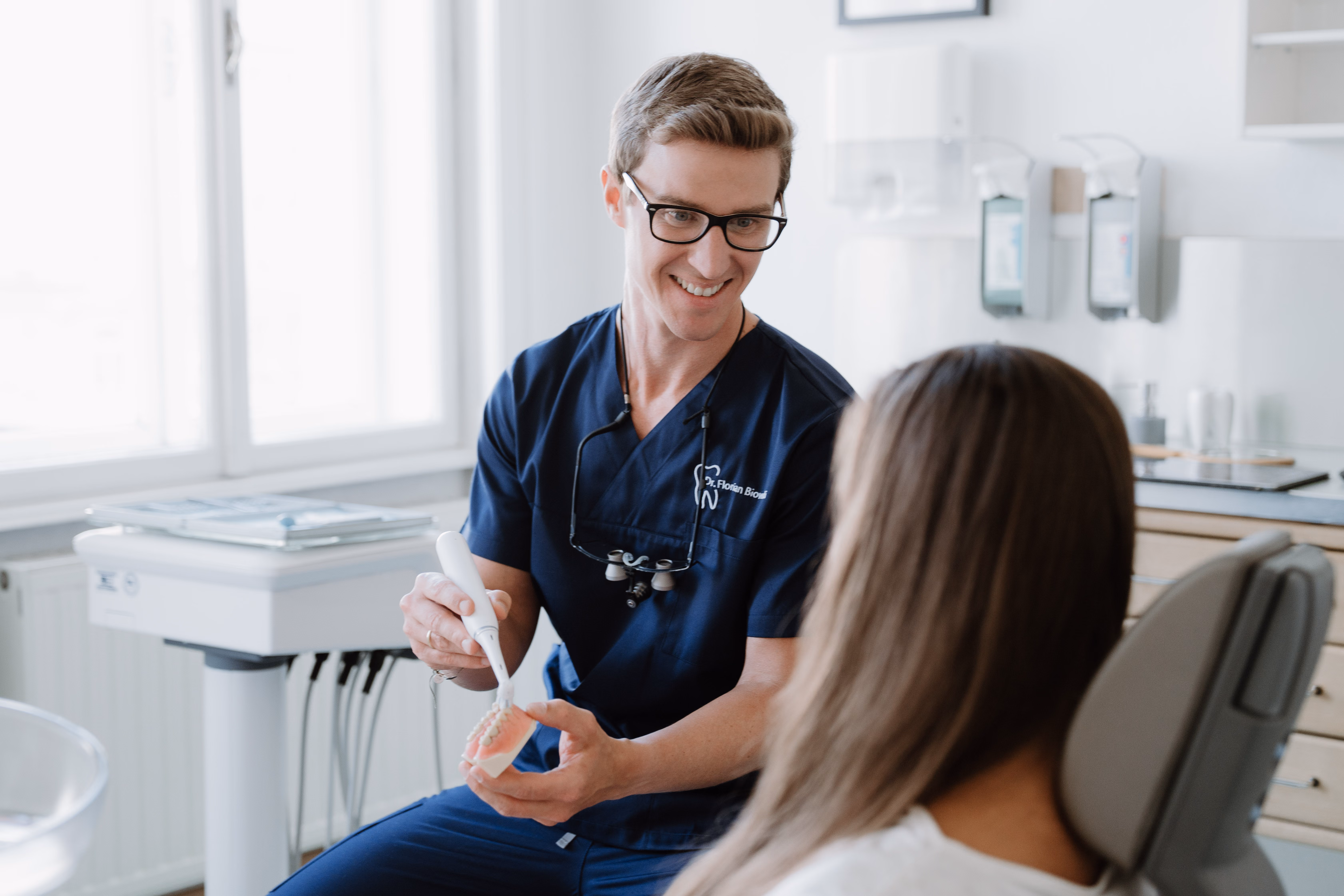 Zahnarzt erklärt Patientin die Zahnreinigung mit einem Zahnmodell und einer Bürste.