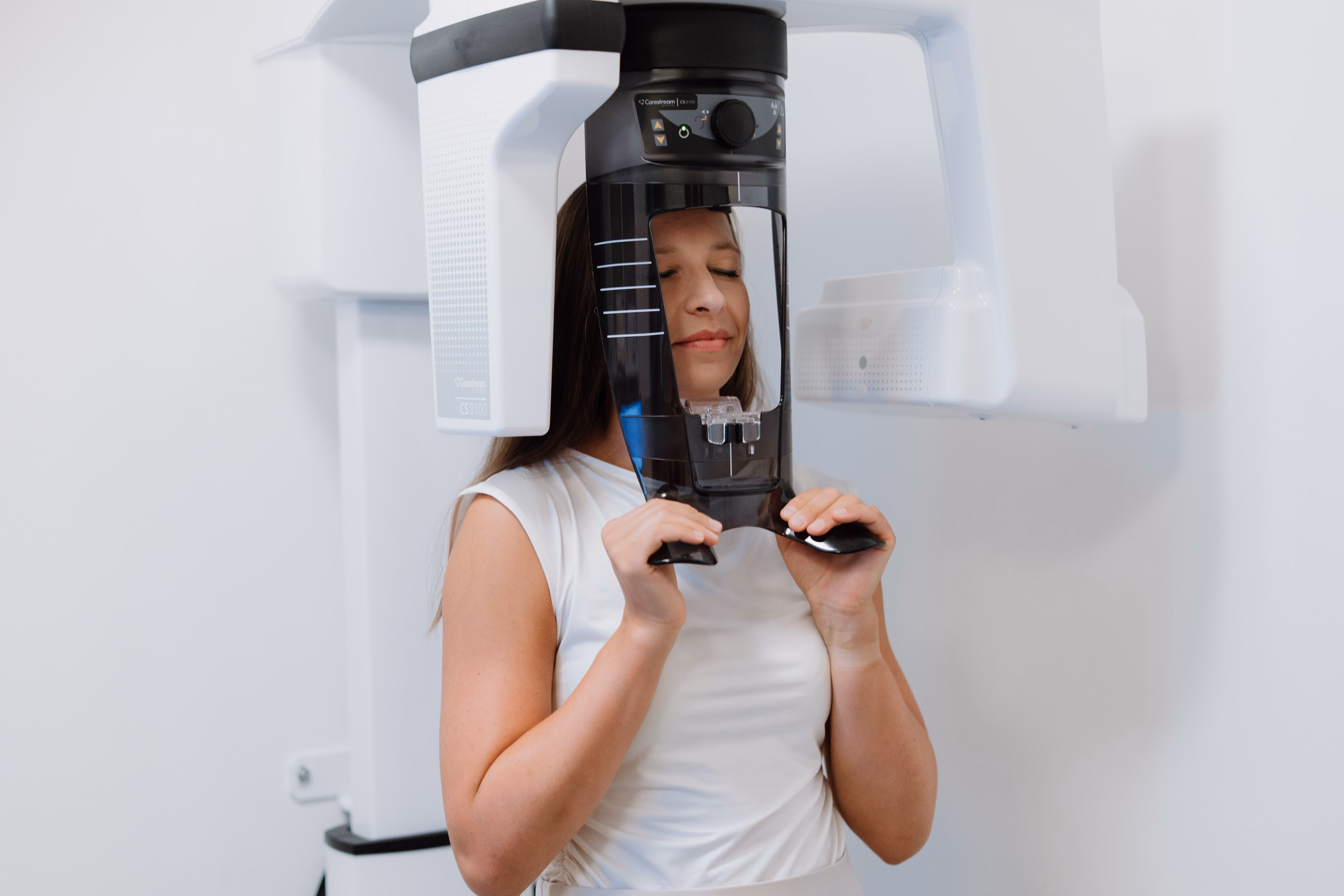 Frau in weißem Top bei einer zahnärztlichen 3D-Röntgenaufnahme mit geschlossenem Gesichtsausdruck.