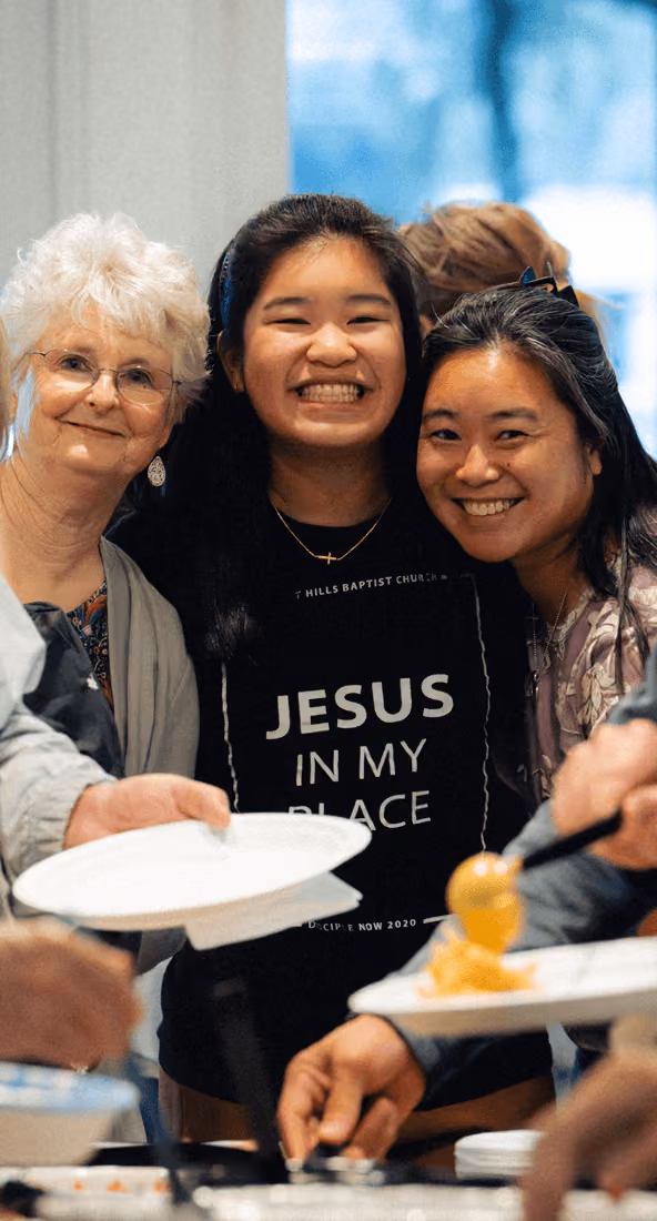 Three smiling women at a gathering, one wearing a shirt that says 'Jesus in my place'.
