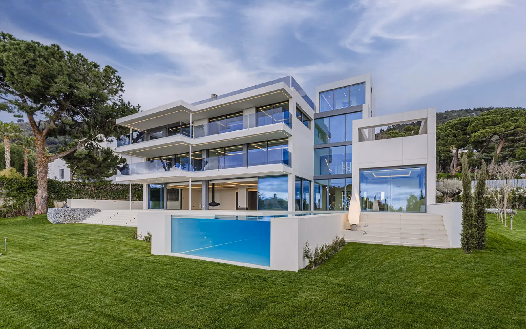 Modern three-story house with glass balconies, large windows, and a transparent pool wall on green lawn.