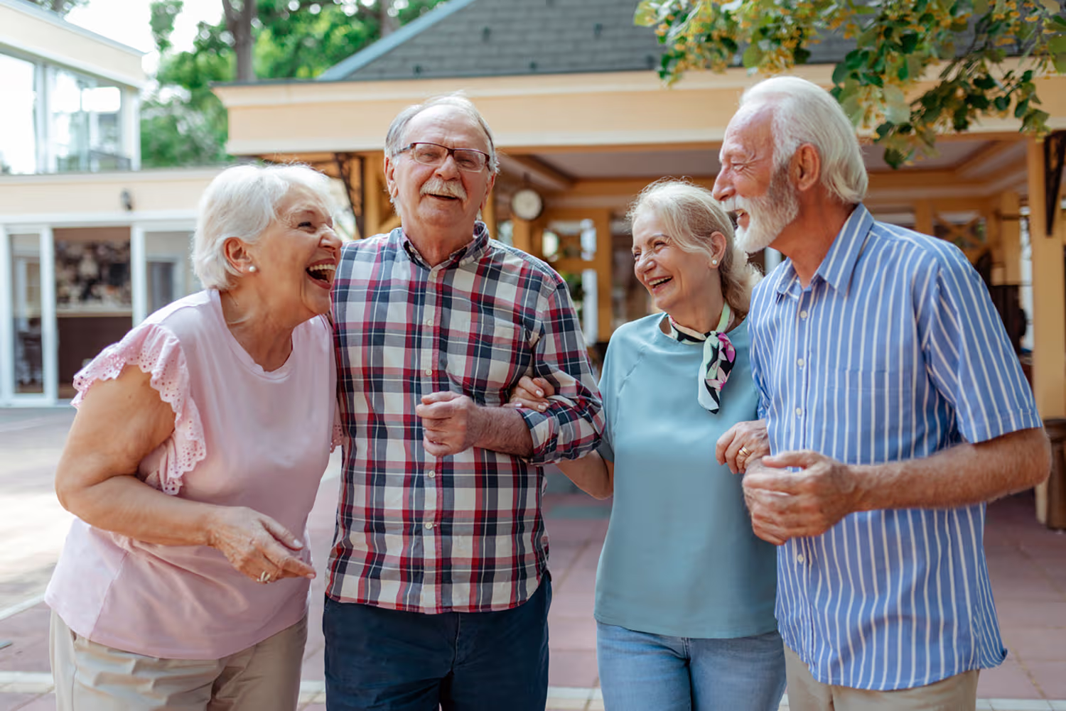 Four elderly friends laughing and walking arm in arm outdoors near a building.
