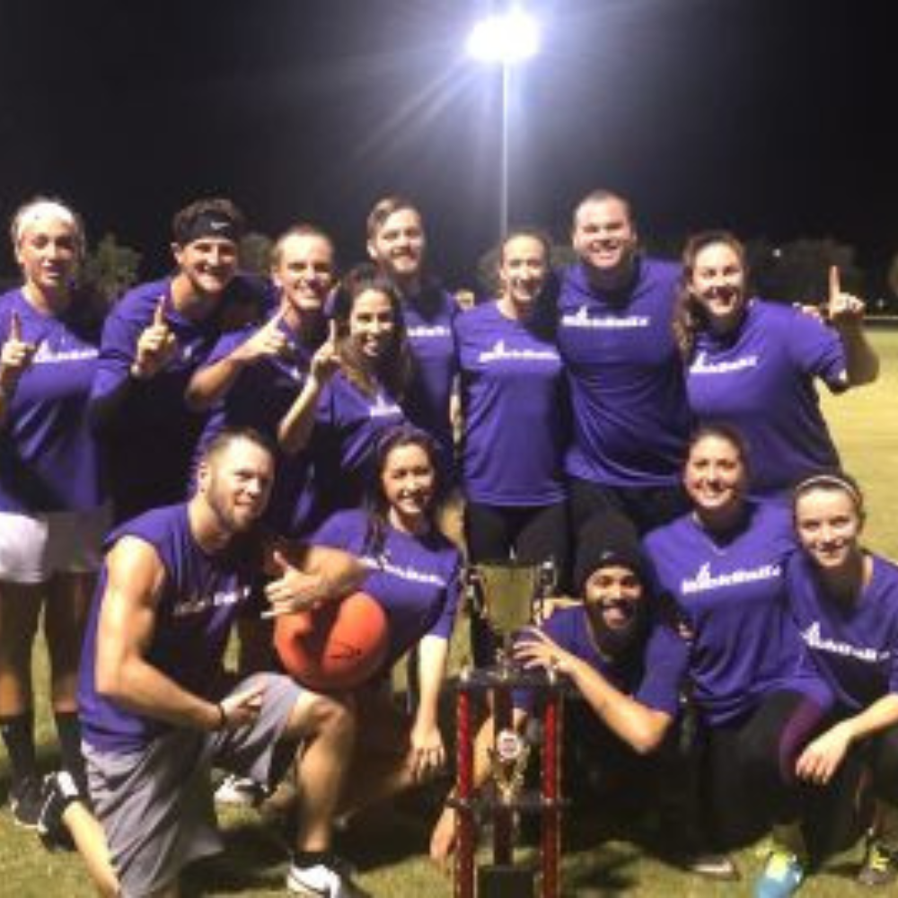 Group of athletes in purple shirts posing with a trophy and a ball on a lit outdoor field at night.