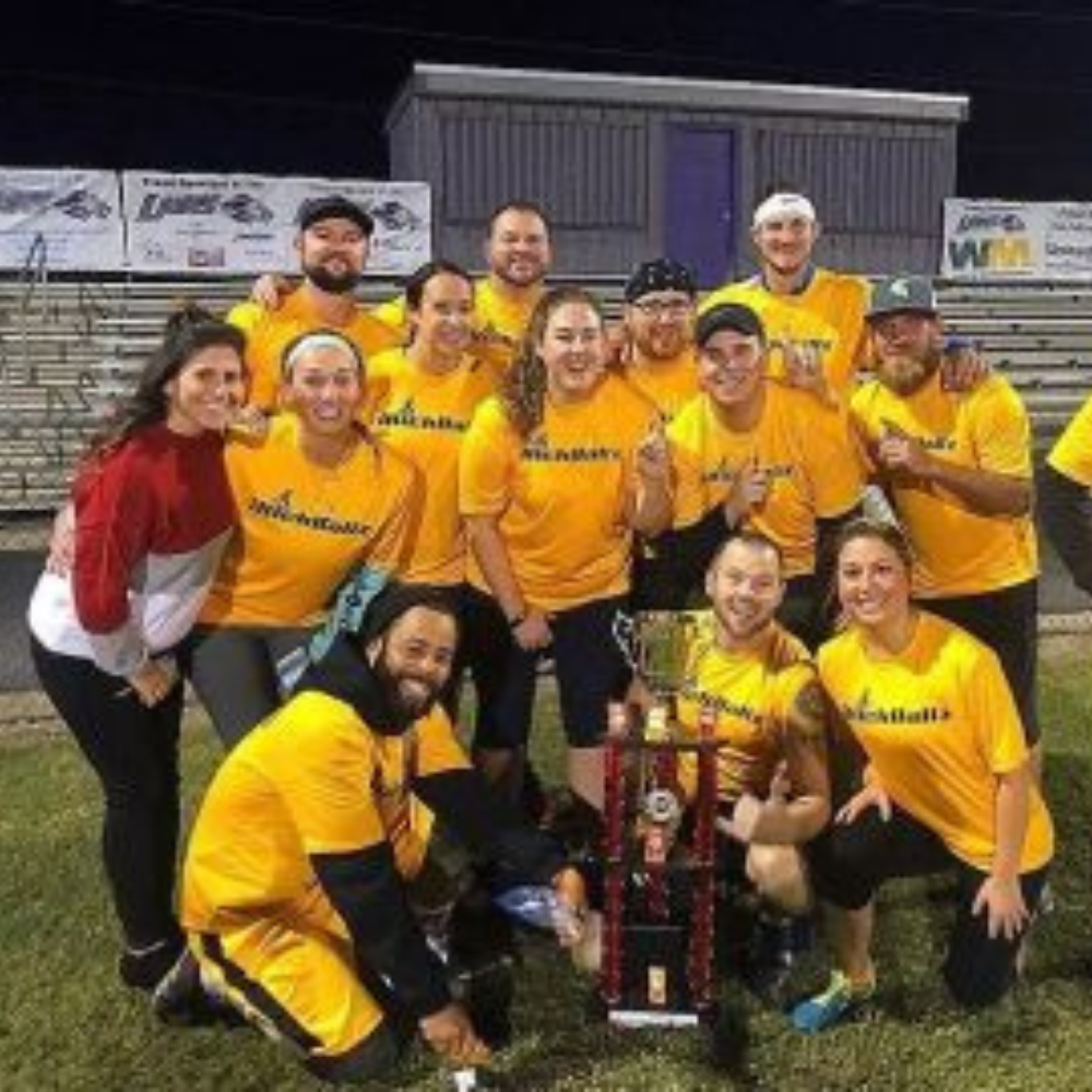 Group of people in yellow shirts posing on field at night, holding a large trophy and smiling.