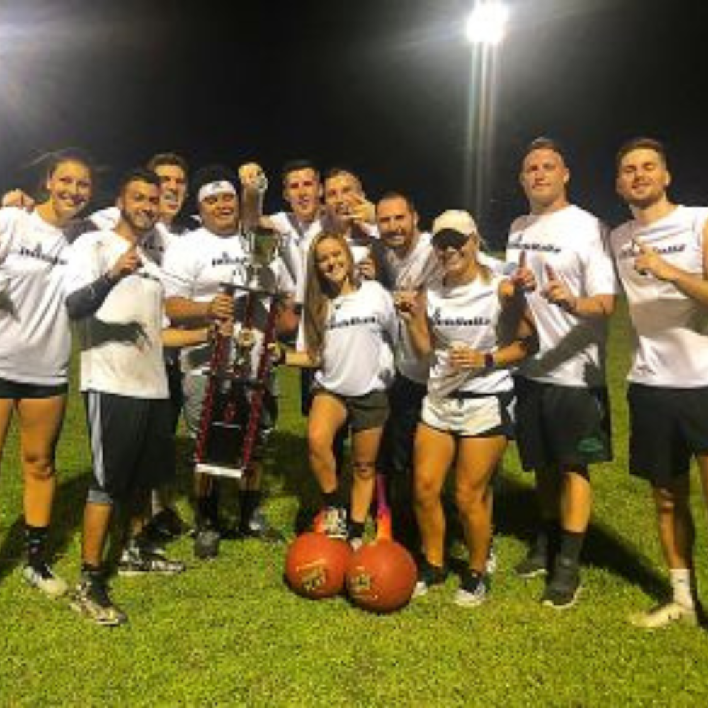 Group of people in white shirts posing on grass at night with a large trophy and two red medicine balls.