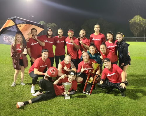 Group of people wearing red shirts posing on grass with trophies and an umbrella at night.