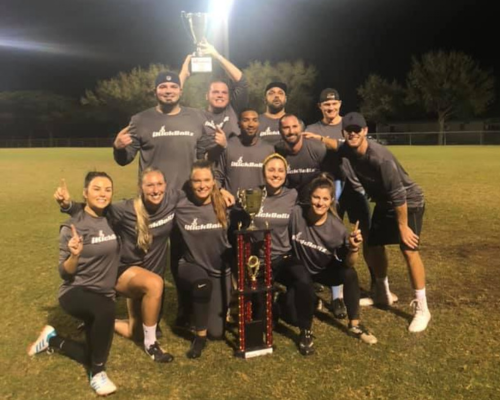 Group of ten people posing on grass at night with a large trophy and a small trophy, smiling and showing number ones.