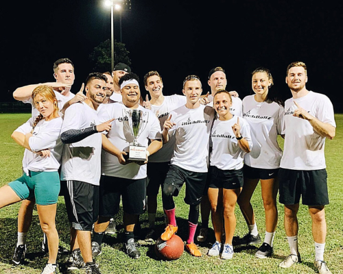 Group of people posing on a field under lights, holding a trophy and celebrating a sports win.