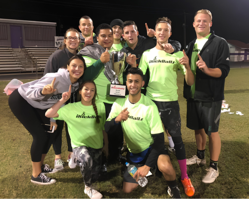 Group of nine people in green shirts holding a large trophy on a sports field at night.