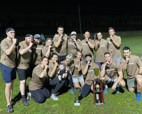 Group of 15 people posing outdoors on grass with a trophy and a red ball, many wearing matching pink jerseys.