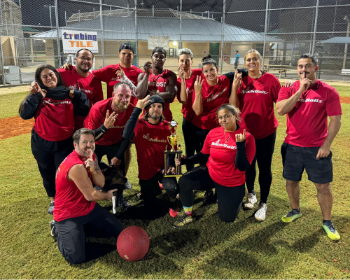 Group of people wearing red shirts posing on grass with trophies and an umbrella at night.