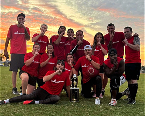 Group of ten people posing on grass at night with a large trophy and a small trophy, smiling and showing number ones.