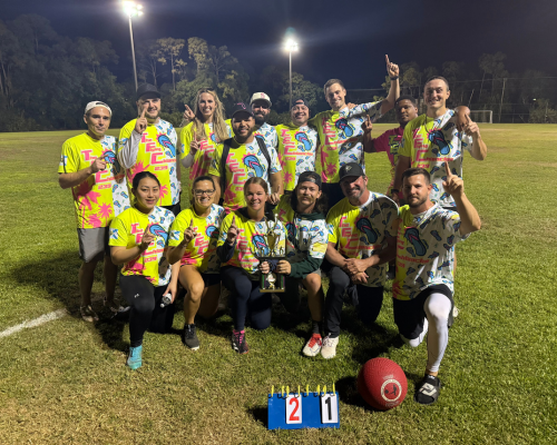 Group of 15 people posing outdoors on grass with a trophy and a red ball, many wearing matching pink jerseys.