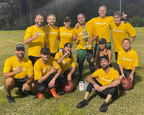 Group of people in blue iKickBallz shirts posing outdoors at night on grass field.