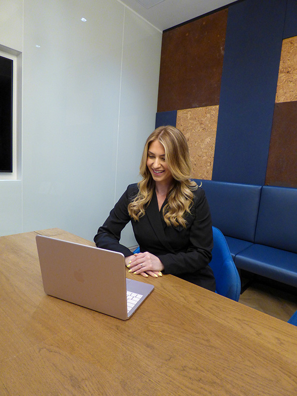 Smiling woman with long blonde hair sitting at a table and using a silver laptop in a modern office.