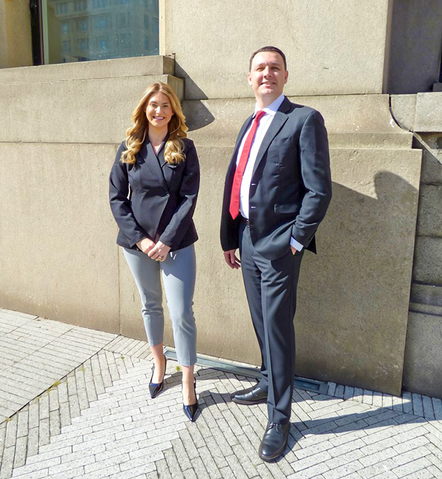 Man in dark suit with red tie and woman in black blazer and gray pants standing outside on a sidewalk.