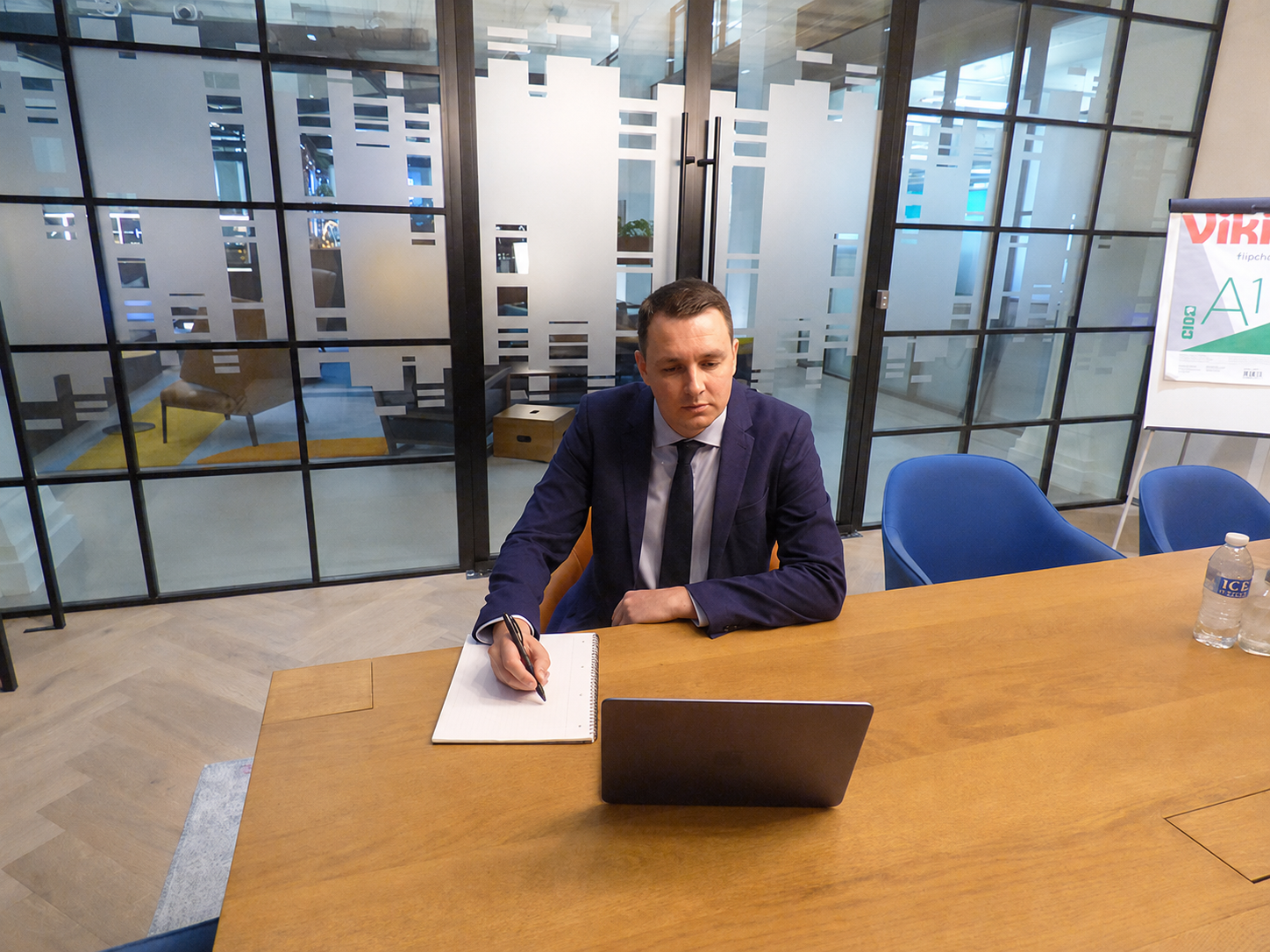 Man in suit working at a wooden table with laptop and notebook in a modern office meeting room.