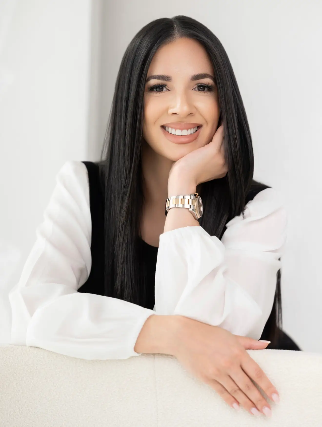 Smiling woman with long black hair resting her chin on her hand wearing a white blouse and watch.