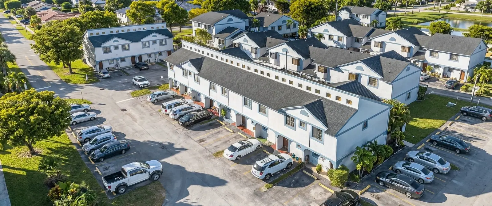Aerial view of a residential complex with white two-story townhouses and parked cars in a sunny area.