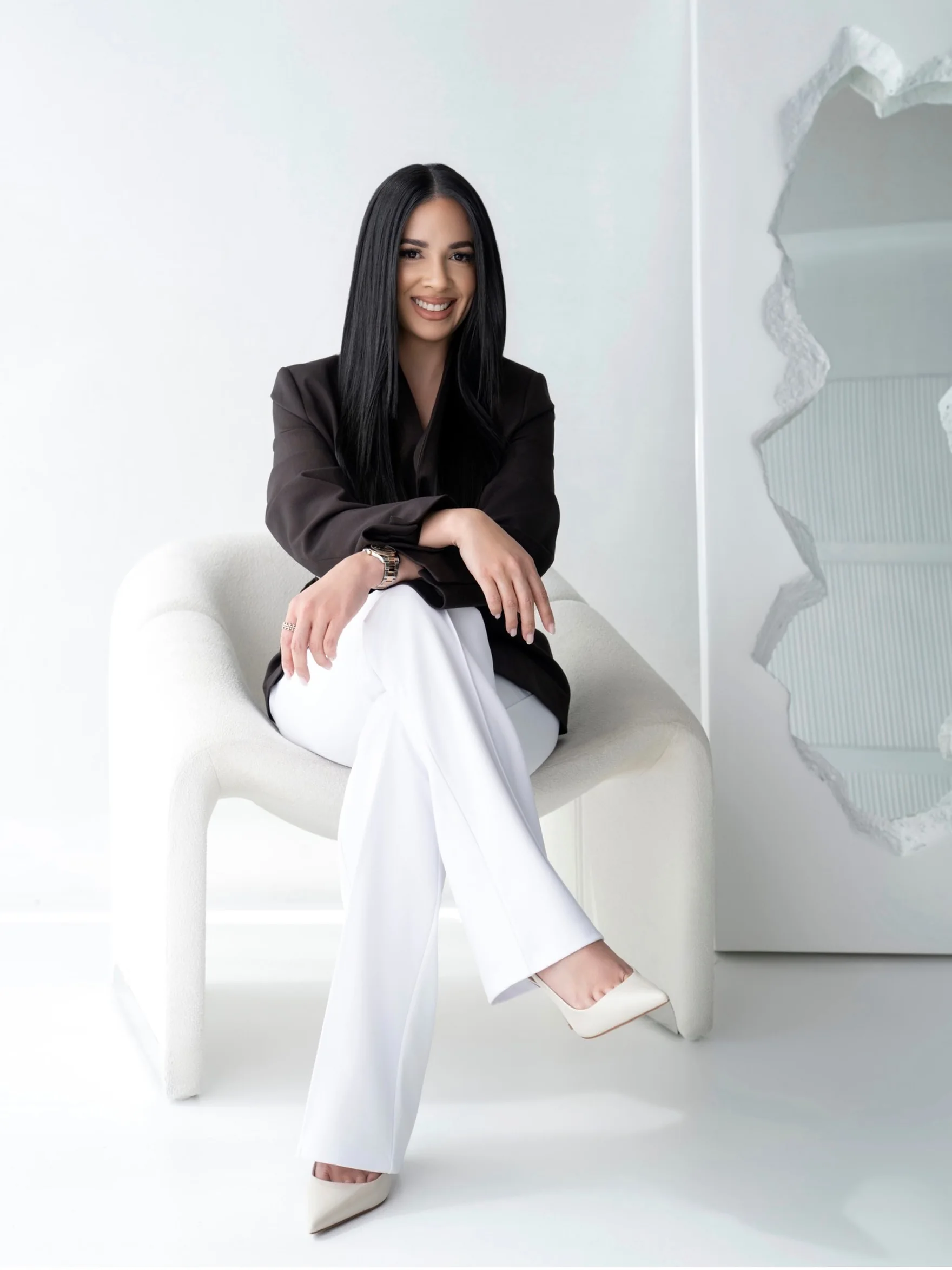 Smiling woman with long black hair wearing a black blazer and white pants sitting on a white chair.