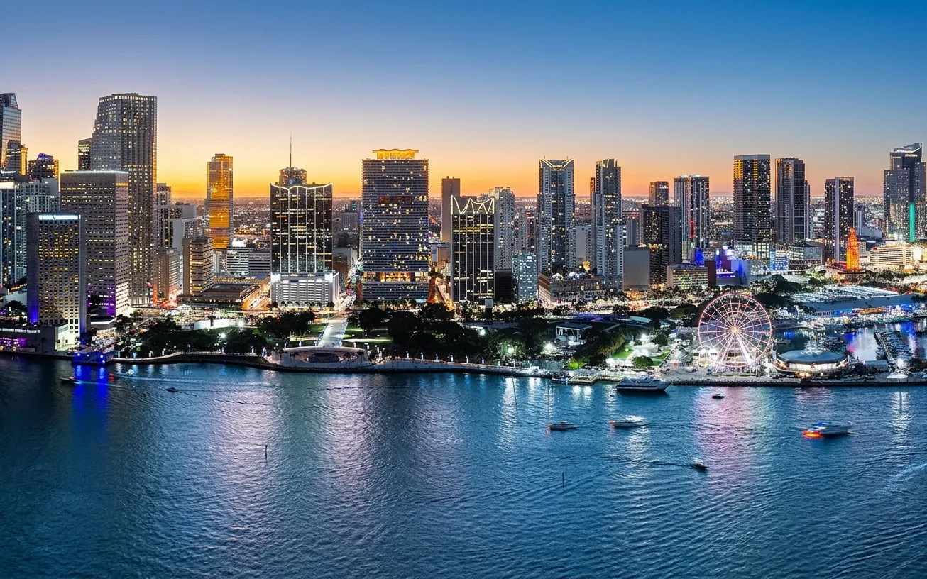 Night view of a city skyline with illuminated skyscrapers and a lit Ferris wheel by the water.