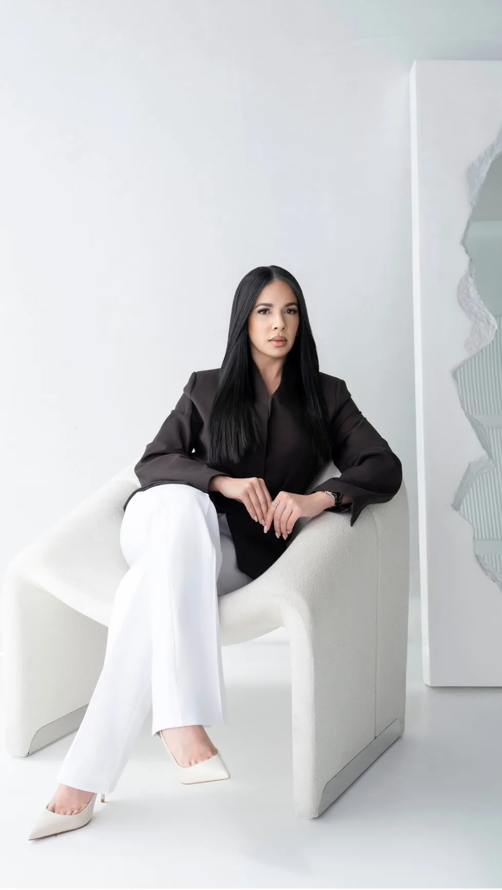 Woman with long black hair wearing black jacket and white pants, sitting in a white chair.